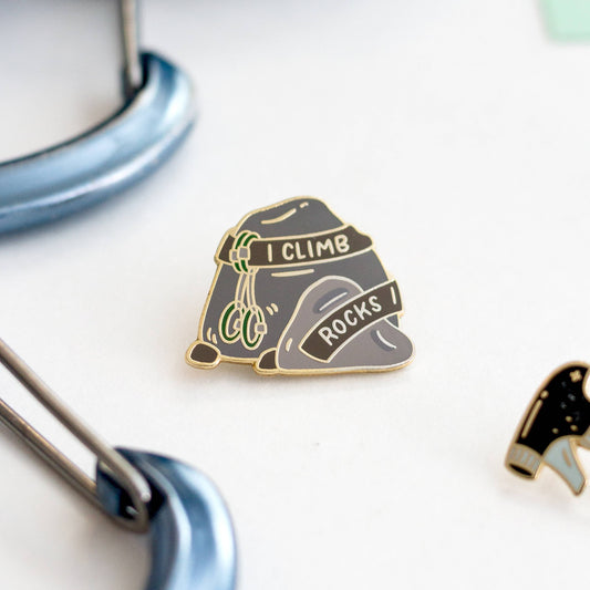 I Climb Rocks Enamel Pin shaped like rocks, displayed next to climbing gear and a small pin.