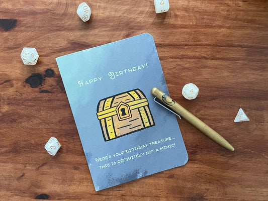Not A Mimic Birthday Card featuring a treasure chest, dice, pen, and Happy Birthday text.