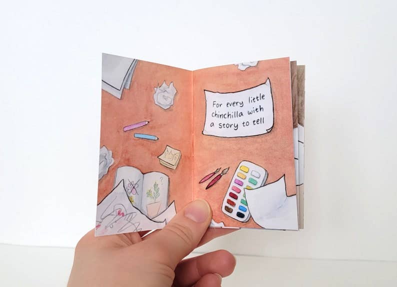 The Chinchillustrator Mini-Comic Zine held open in a hand, with visible art supplies and the text For every little chinchilla.