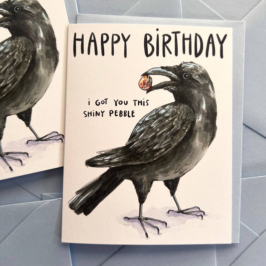 Crow I Got You This Shiny Pebble birthday card featuring a watercolor crow holding a pebble in its beak.