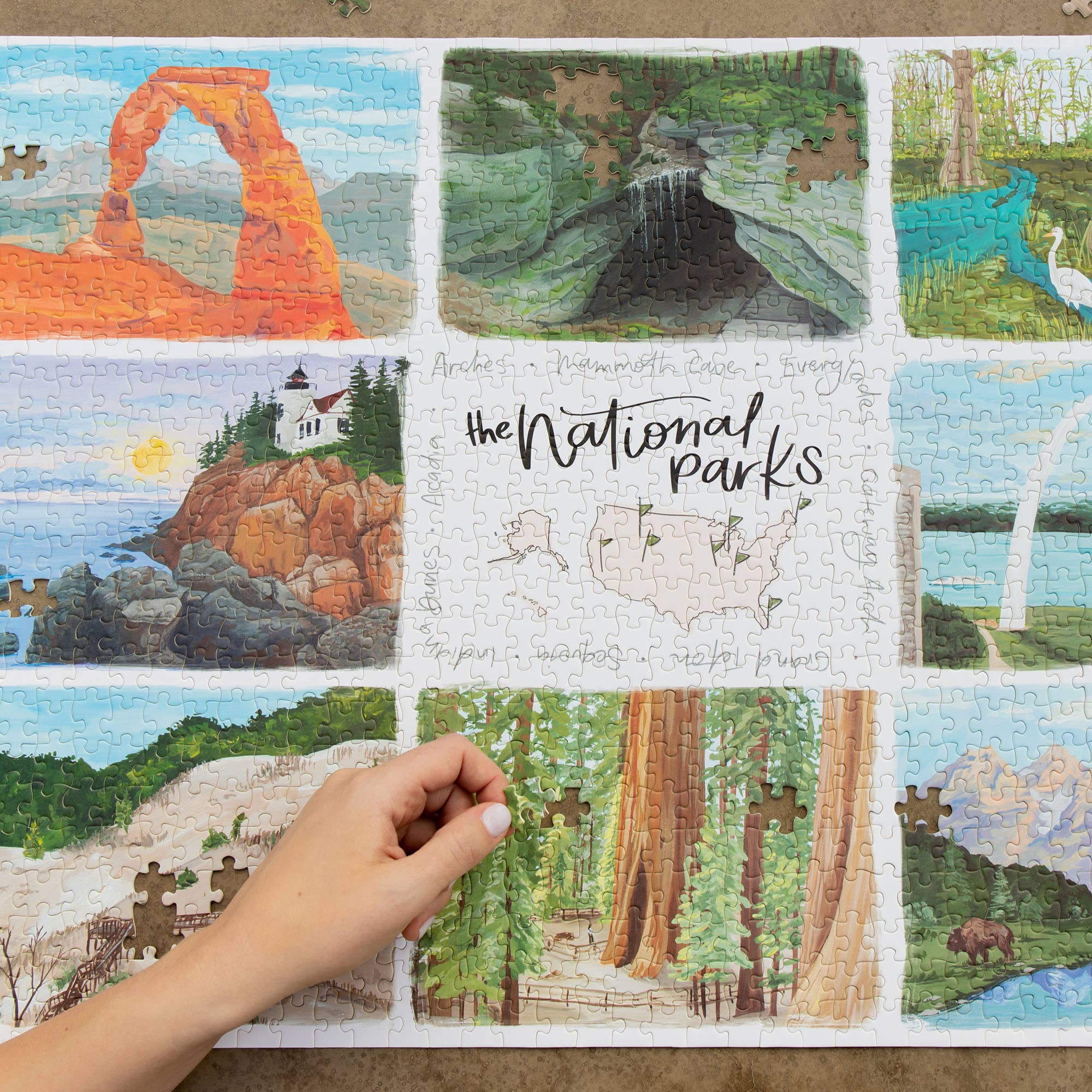 National Parks 1,000 piece jigsaw puzzle with scenic park illustrations, hand placing a piece.