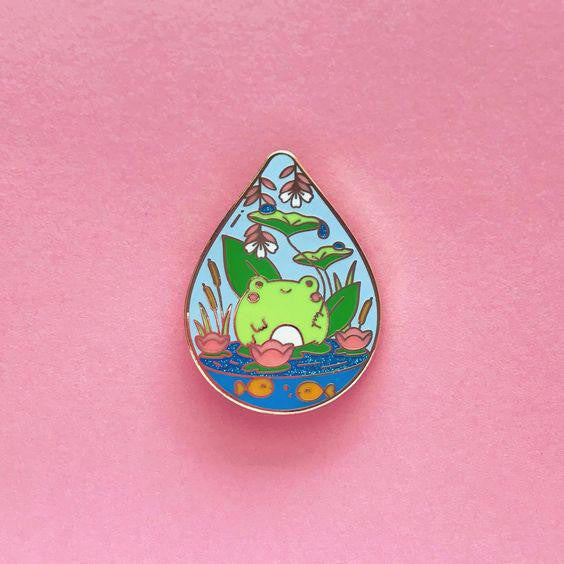 1.5 glitter enamel pin featuring a green frog in a pond with lilies and flowers on a pink background.