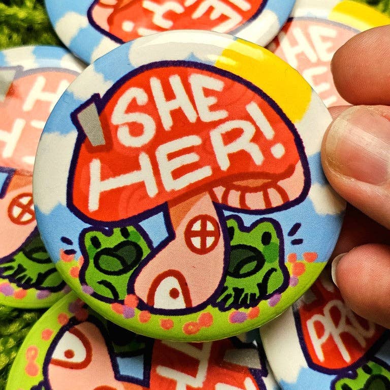 Large laminated waterproof button with Pronoun Frog Mushroom design and SHE/HER text, held between two fingers.