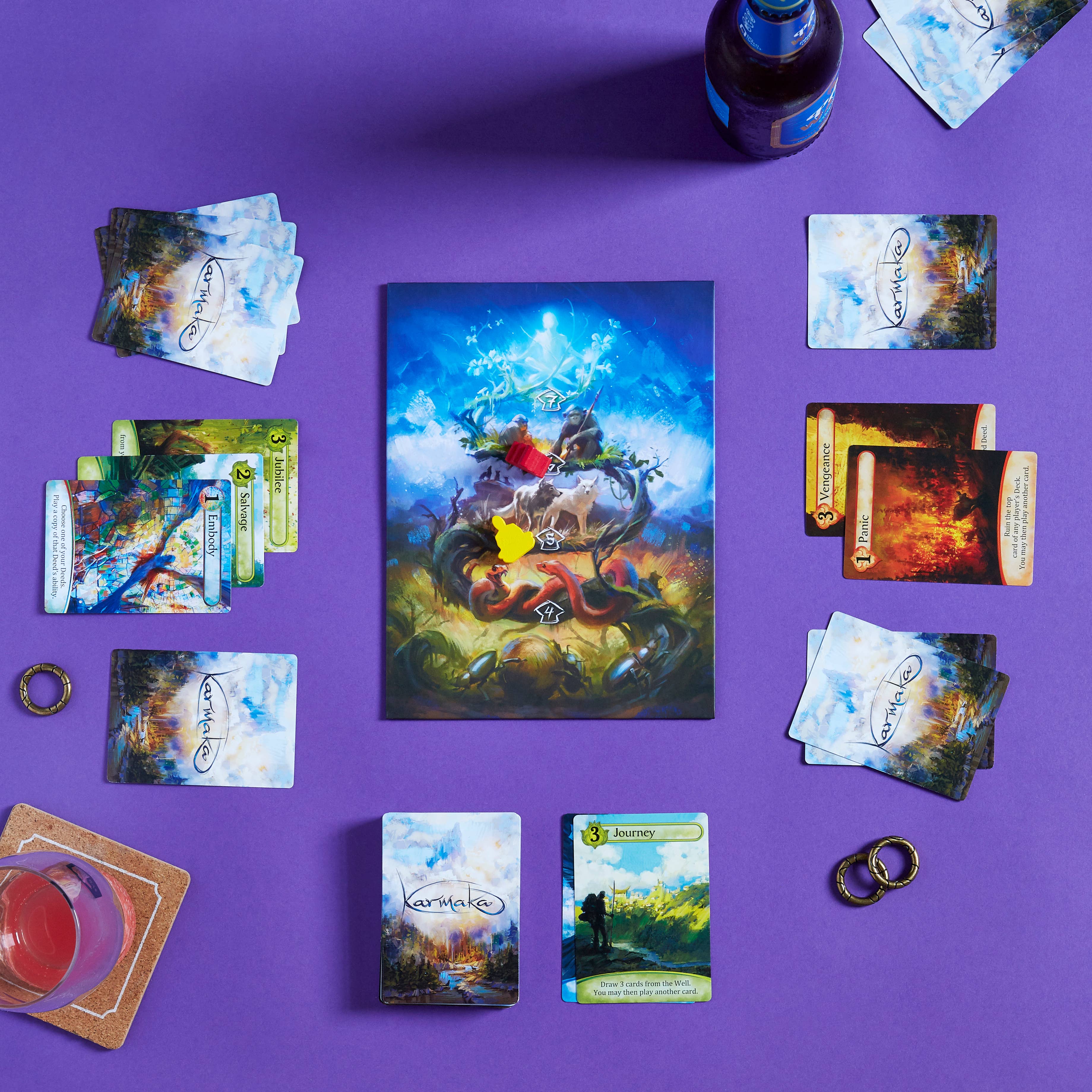 Karmaka card game with cards and tokens arranged on a purple surface; drinks shown nearby.