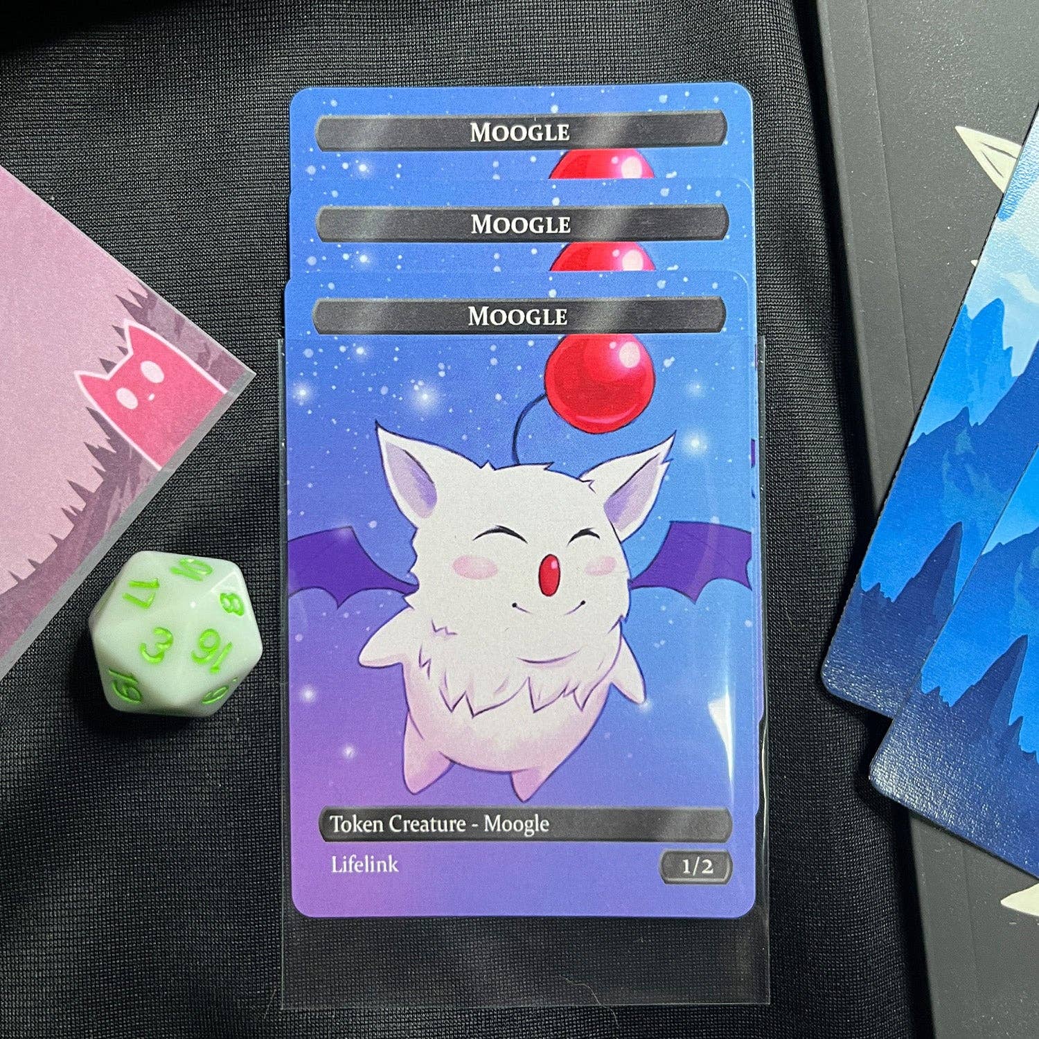 Four Moogle hero tokens displayed on a playmat with green d20, blue Magic cards, and card sleeves.