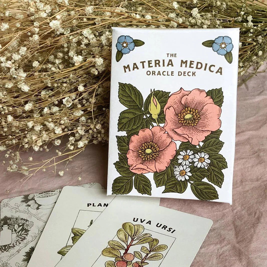 The Materia Medica Oracle Deck with illustrated cards displayed on a pink cloth beside dried white flowers.