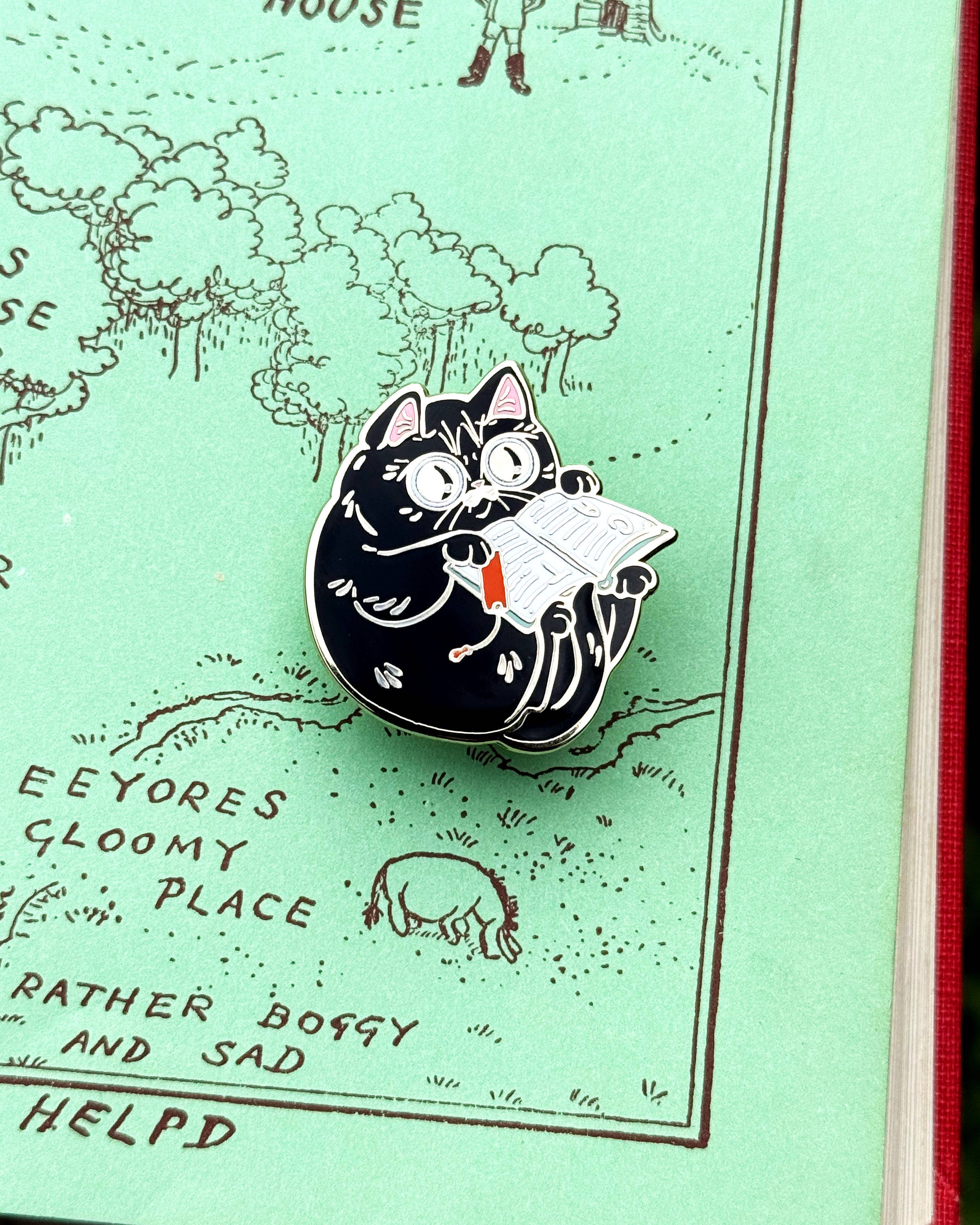 Round cat enamel pin reading an open book with a red bookmark.