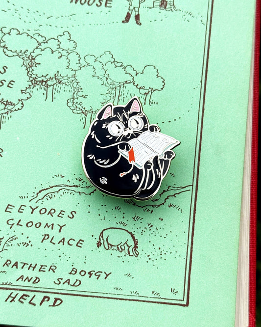 Round cat enamel pin reading an open book with a red bookmark.