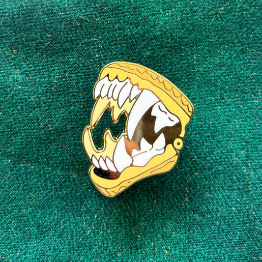 D&D Class Curiosities gold enamel pin shaped like an animal jaw with sharp teeth, shown on a green textured background.