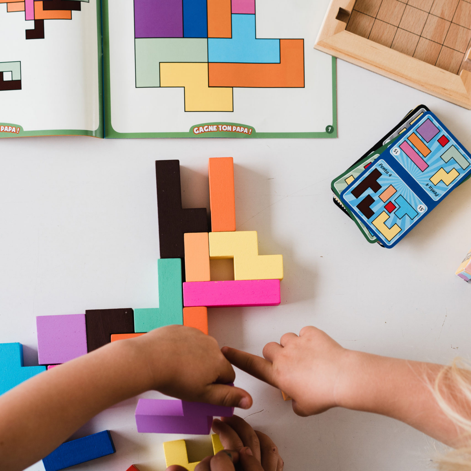 Katamino Family wooden puzzle game with children’s hands assembling pieces on a table; books are nearby.
