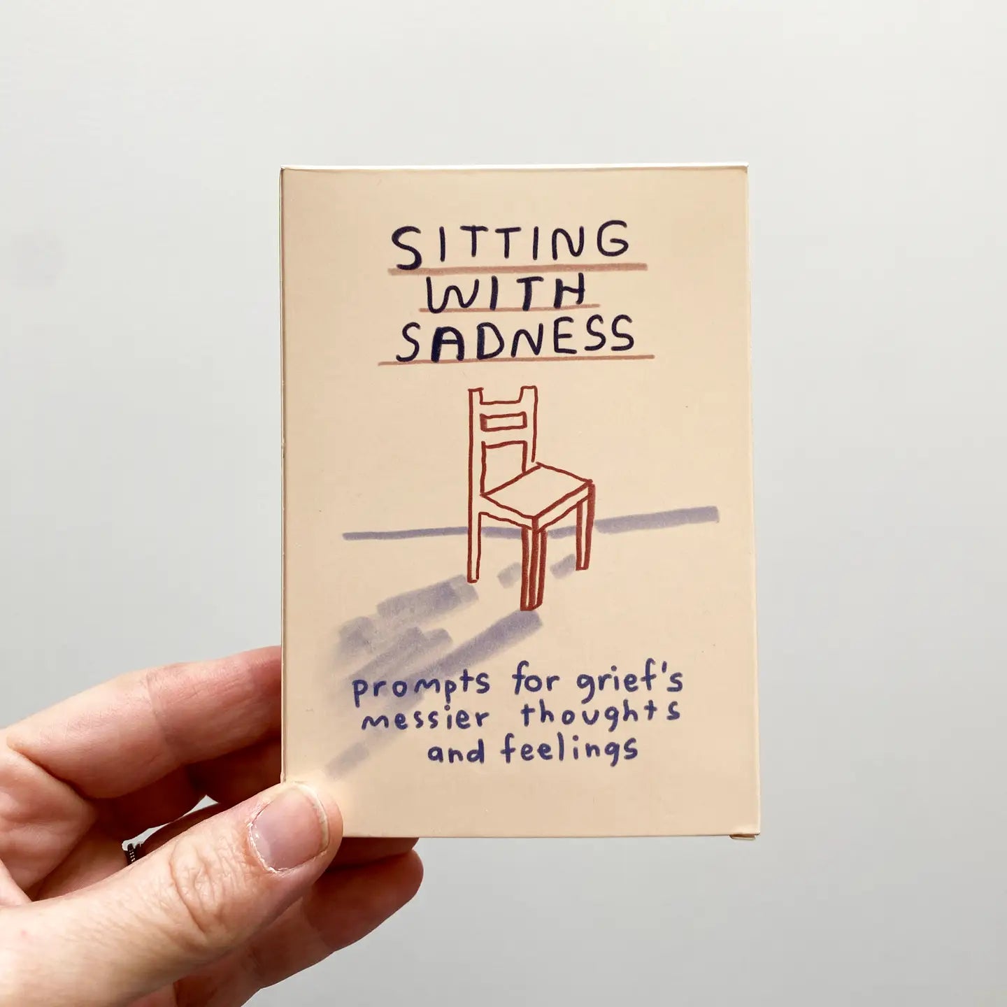 Sitting With Sadness Deck held in one hand, showing a card with a chair and handwritten grief journal prompts.